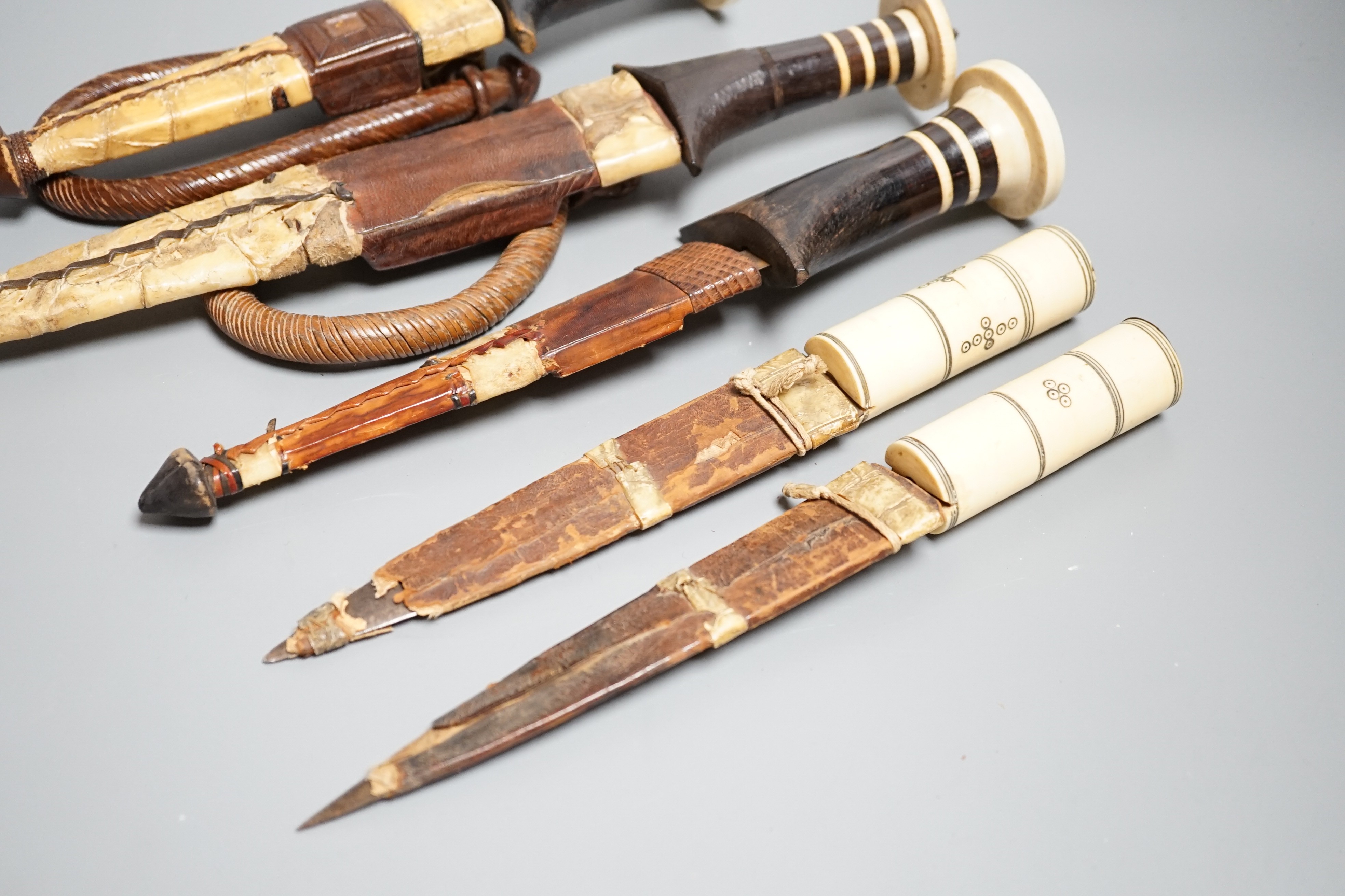 Five assorted African Nilotic tribal daggers c.1900, comprising three with turned ivory mounted ebony handles (two with silver pommels); and two further with cylindrical ivory handles, all in their leather sheaths, maxim